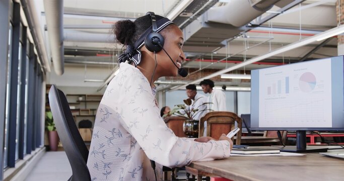 Working woman wearing headset and patterned blouse, checking smartphone and charts at open office