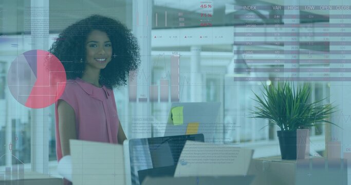 Standing analyst in sleeveless pink blouse pausing at desk in office, with laptop monitor notes