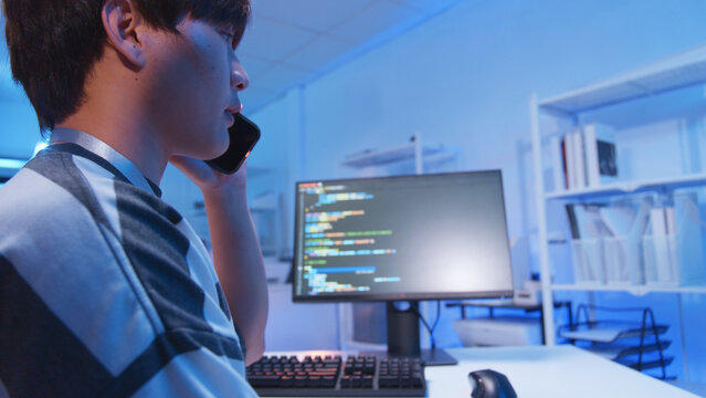 Young male Asian IT freelancer programmer and tech engineer remotely works with computer and mobile phone, software codes for application development, and web network system information data in room.
