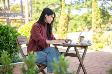 Asian woman typing on laptop aside coffee and headphone while sitting at wooden table in garden cafe