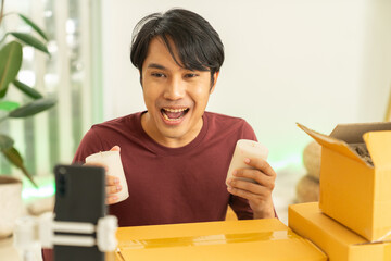 Young man selling online holding candles excitedly showing products for ecommerce business at home with boxes and smartphone camera