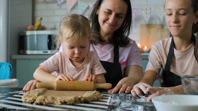 A young girl rolls ginger cookie dough under the gentle supervision of her mother, while the older sister assists with baking tasks nearby, family cooking moment concept