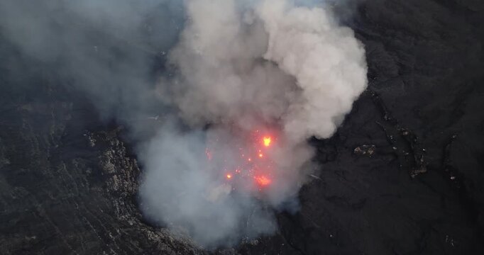 Mount Yasur volcano erupting from an aerial drone viewpoint, showing molten lava explosions and thick smoke rising from the active crater in a dramatic display of raw power and natural hazard