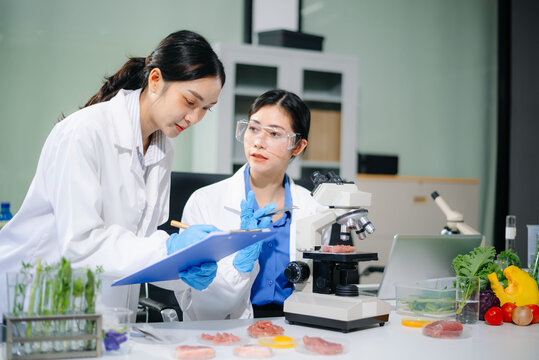 Asian food scientists inspect raw meat samples under microscope for biotech research, hygiene, and food safety analysis.