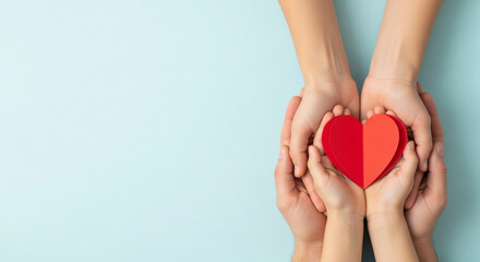 Top view of multiple pairs of hands, adult and child, gently cupping and protecting a bright red paper heart against a light blue background with copy space