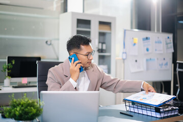 Asian businessman talking on smartphone while working on laptop at modern office desk, symbolizing business communication and corporate success.