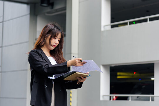 Young Asian business woman in suit busy with work, reviewing important documents and taking notes on city street holding pen and notebook, professional task demonstrating dedicated employee