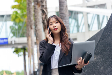 Young Asian woman, business professional, work suit clad, busy on street talking phone, holding laptop, managing tasks in modern urban city environment, dedicated to career success