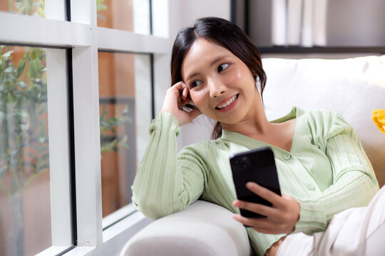 Young asian businesswoman looking smartphone smiling and relax sitting on sofa in living room at home, cheerful woman watching smart phone sitting on couch in home, lifestyle and communication.