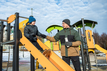 A father helps his child down a bright yellow children's slide in a snowy city park, both dressed in winter jackets, hats and gloves.