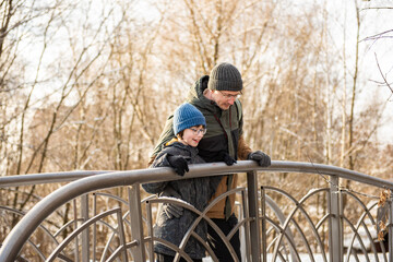 A father and son are standing together on a small pedestrian bridge in a snowy park, leaning on the railing and looking at the winter landscape.