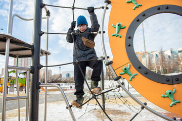 A child climbs a rope net on a colorful outdoor playground in a snowy urban park, wearing a blue beanie, winter coat and a small crossbody bag.