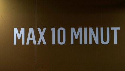 Sign “MAX 10 MINUT” on dark parking wall with partial car silhouettes, highlighting time restriction, urban signage, minimal composition, and controlled space atmosphere.