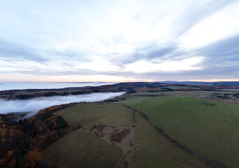 Aerial view of wide fields under dramatic cloudy sky with low mist hugging forest edge, highlighting serene atmosphere, natural depth, and expansive landscape horizon.