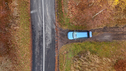 Top-down aerial view of blue car parked by asphalt road on dirt turnout amidst autumn foliage, capturing contrast of natural colors, vehicle presence, and quiet roadside moment.