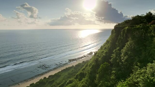 Lush green cliffside overlooking a serene sandy beach along the coastline of Bali, with the sun low on the horizon casting a brilliant golden reflection across the tranquil ocean waters