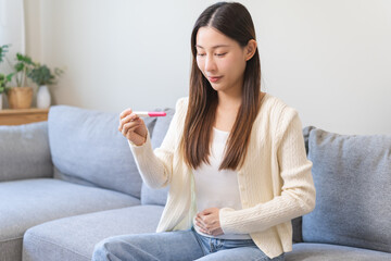 Happy thankful asian young woman hand holding pregnancy test after result with positive, surprised glad girl sitting on couch living room at home. Pregnant of ovulation fertility, motherhood concept.