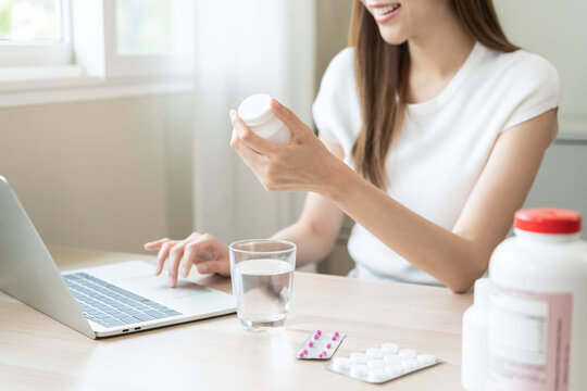 Wellness and dieting asian young woman, girl working from home using computer, typing or searching prescription on medicine label about vitamins information online, holding capsules of food supplement