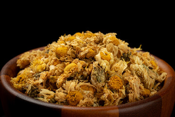 Pile of dried chrysanthemum flowers in a brown wooden bowl on black background.
