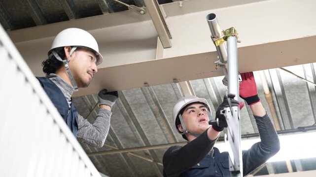 天井が近い高所の足場で単管パイプを固定するクランプなどの施工作業の動画　鳶職や作業員のイメージ　作業進捗を確認する2人
