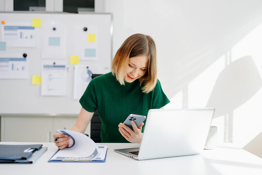 Business woman reviewing documents while checking her smartphone at a bright modern desk.