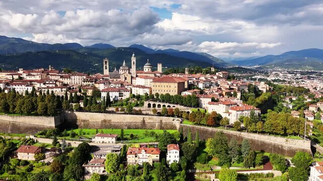 A panoramic view of Bergamo, Italy features terracotta rooftops, medieval towers, fortified walls and lush greenery set against rolling hills and distant mountains under dynamic clouds daylight today