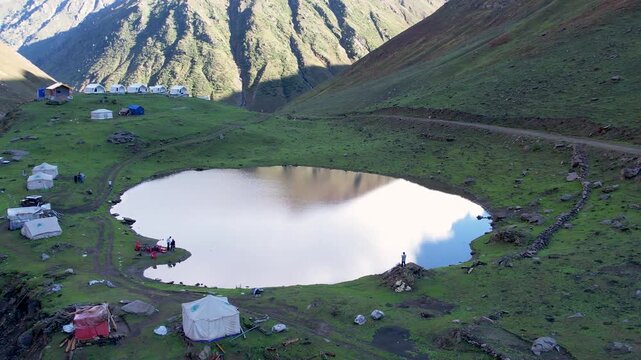 Tents around Chinar Lake in Neelum Valley, reflecting sky amid green slopes and rugged terrain. Kashmir, Pakistan