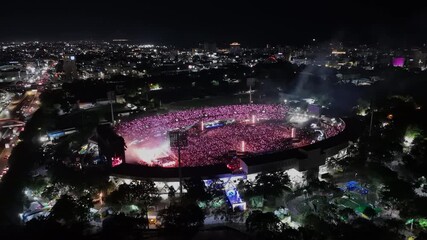 Aerial approaching shot showing crowd of people celebrating bad bunny festival show in open air stadium at night. Santo Domingo, Dominican Republic. Music show at night with flashing illuminated light