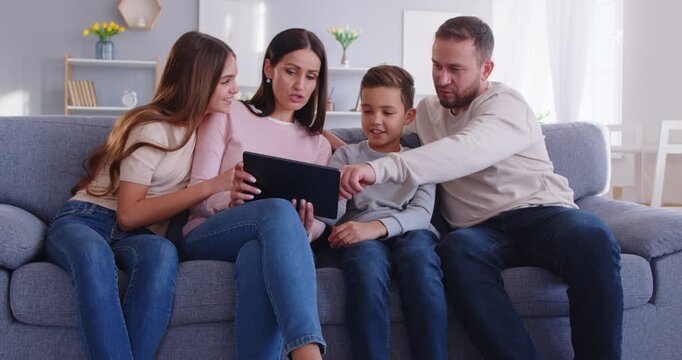 Family of four members sitting at sofa using tablet and internet connection for doing shopping online choosing new furniture for home discussing something. Gadget help in family issues