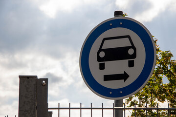 Circular traffic sign indicating right turn for vehicles, featuring a simple car icon, set against a cloudy sky with greenery in the background, emphasizing road safety and navigation