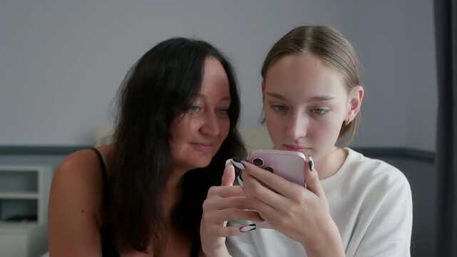 Woman with long dark hair in black sleeveless top sitting beside blonde teenage girl in white t-shirt, both intently viewing pink smartphone held by teenager against neutral gray wall background.