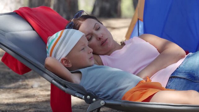 A mother and her son lie together on a grey camping cot. They relax and smile near a blue tent in a sunny pine forest during a family holiday.