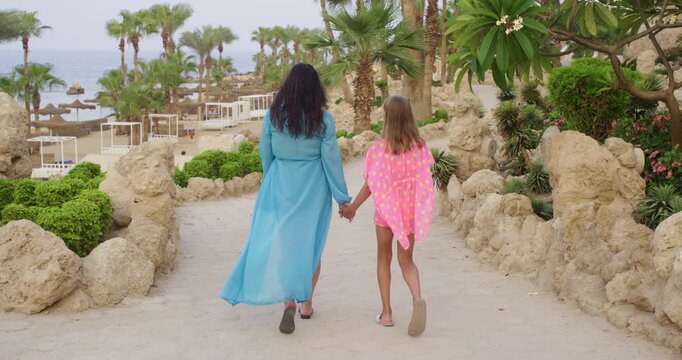 Woman in turquoise blue maxi dress and blonde teenage girl in pink patterned cover up walking together holding hands on sandy path with palm trees, rocks, beach loungers, and sea in background.