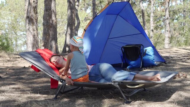 A mother and her son lie together on a grey camping cot. They relax and smile near a blue tent in a sunny pine forest during a family holiday.