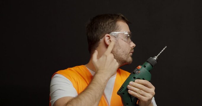 A worker inserts earplugs and points to his ear while holding a drill, emphasizing hearing protection and safety against loud noise at work.