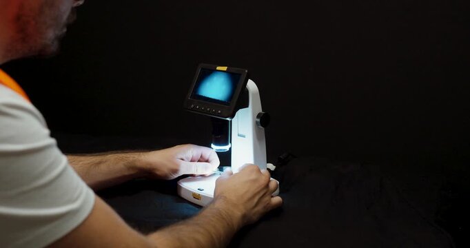 A person operates a digital microscope under focused light, examining a small object on the display for detailed inspection and precision work.