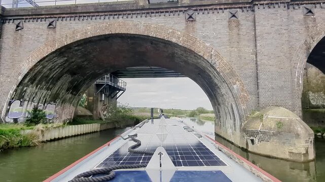 View from the stern of a narrowboat cruising under a set of old and modern railway bridges on the river Nene in Northamptonshire, England.