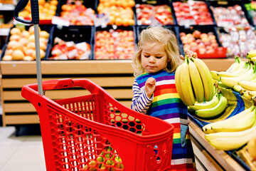 Cute todler girl pushing shopping cart in supermarket. Little child buying fruits. Kid grocery...