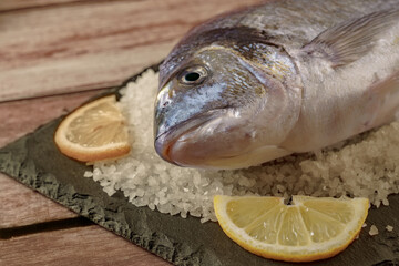 Whole fresh sea bream uncooked on a slate board with coarse sea salt and lemon slices, ready for cooking