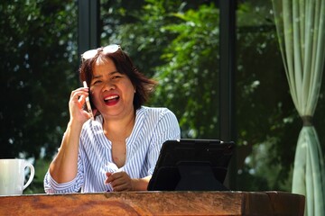 A woman is sitting, talking on a mobile phone and laughing happily, with a tablet on the table by the window.