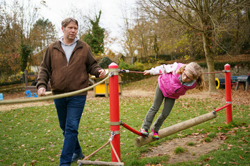 Little school girl playing with father on outdoor playground. Happy child having fun with climbing, swinging. Dad help daughter.
