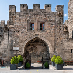 Kayseri Castle, an ancient stone fortress with an arched entrance, barred windows, and battlements, stands under a clear sky in Kayseri, Turkey. Potted plants flank the entrance