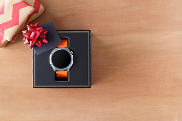 Top view of a smartwatch with an orange band in a premium black gift box, with a red bow and a blank card on wooden background. Flat lay with copy space for promotion and christmas, new year gift.