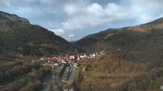 Fast drone approach from afar towards white medieval buildings with orange clay roofing tiles and mountain background amid morning clouds and sun rays.