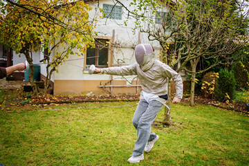 teenage boy practices fencing with a saber, wearing a fencing uniform outdoors.