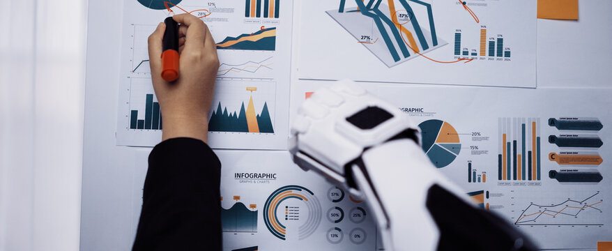 A person is analyzing detailed business infographics while using a robotic arm in a modern office. The scene highlights technology integration in data analysis.Noogenesis