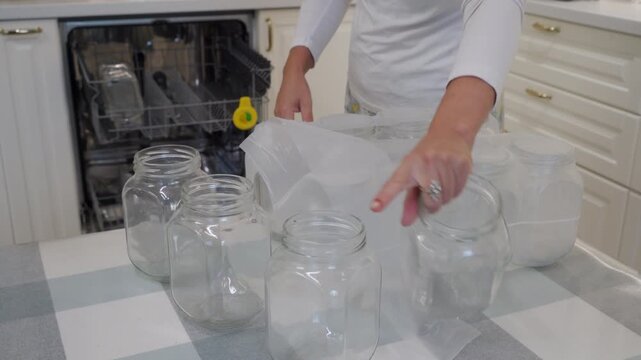 A woman removes new glass jars from plastic packaging at a table. She prepares the containers for canning in her clean kitchen near an open dishwasher.