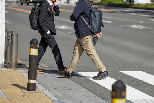 Two businessmen carrying backpacks cross paths on city crosswalk under bright daylight. One is in formal business attire and conversing on smartphone while other is in casual jacket and slacks.