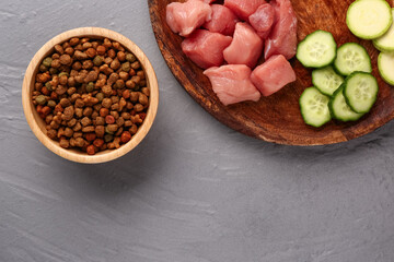 Bowl with dry pet food, raw meat and natural products on color wooden background