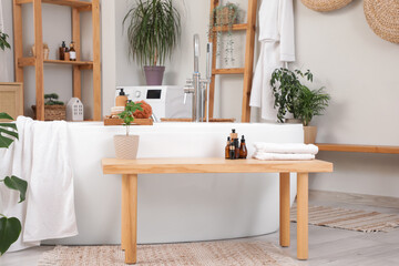 Interior of light bathroom with bathtub and bathing accessories on wooden bench